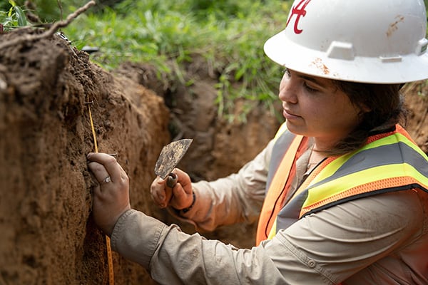 A student wearing a hard hat examines soil in a trench